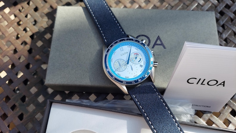 May include: A wristwatch with a blue and white face and a dark blue leather strap. The watch has the brand name "CILOA" on the face and the strap is stitched with white thread. The watch is displayed with its box and instructions.
