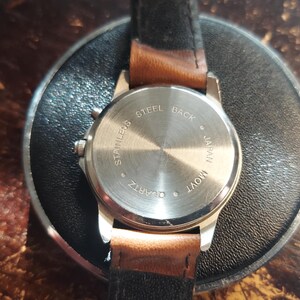 May include: A silver-toned quartz watch with a brown leather strap. The watch back is stamped with "STAINLESS STEEL BACK JAPAN MOV'T QUARTZ". The watch is resting on a black circular surface.