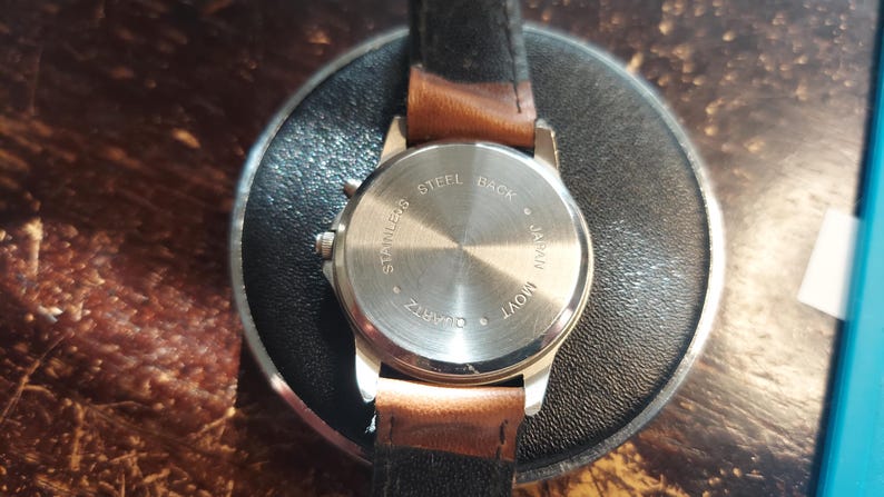 May include: A silver-toned quartz watch with a brown leather strap. The watch back is stamped with "STAINLESS STEEL BACK JAPAN MOV'T QUARTZ". The watch is resting on a black circular surface.