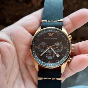 May include: A gold-toned wristwatch with a black leather band. The watch face is dark brown with gold accents, featuring multiple sub-dials and the brand name. The band is stitched with cream-colored thread.