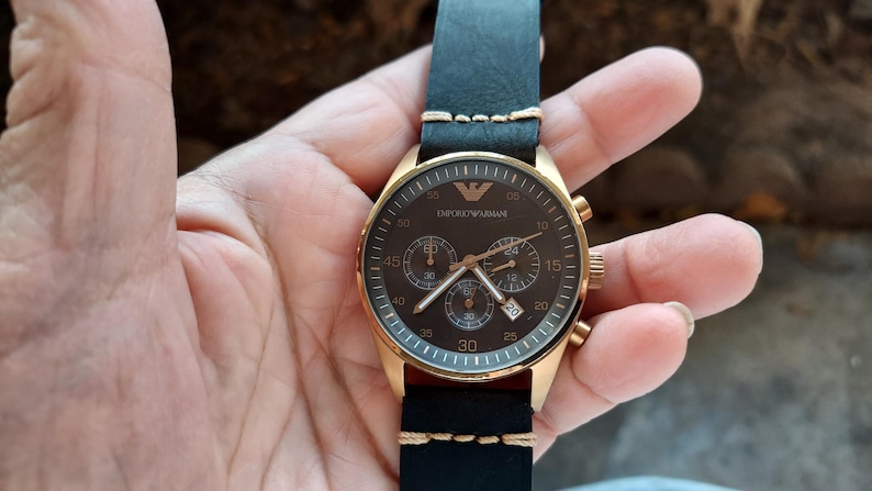 May include: A gold-toned wristwatch with a black leather band. The watch face is dark brown with gold accents, featuring multiple sub-dials and the brand name. The band is stitched with cream-colored thread.