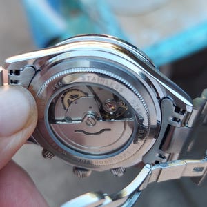 May include: Close-up of a stainless steel wristwatch, showcasing the intricate inner workings through a transparent back. The watch has a silver band and a detailed mechanical movement visible. The watch is held by a person.
