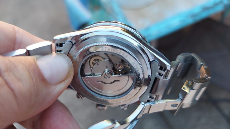 May include: Close-up of a stainless steel wristwatch, showcasing the intricate inner workings through a transparent back. The watch has a silver band and a detailed mechanical movement visible. The watch is held by a person.