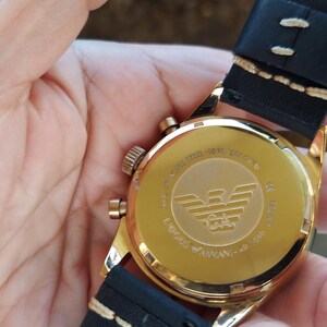 May include: A gold-toned wristwatch with a black leather band. The watch face is visible, displaying the brand logo and text. The watch is held in a person's hand.