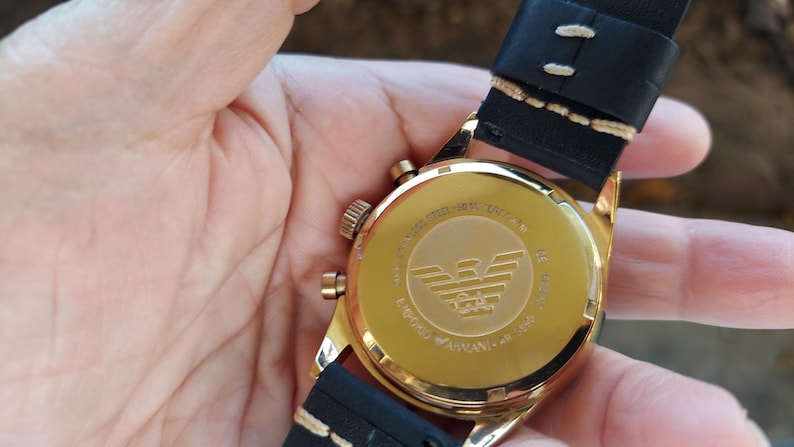 May include: A gold-toned wristwatch with a black leather band. The watch face is visible, displaying the brand logo and text. The watch is held in a person's hand.