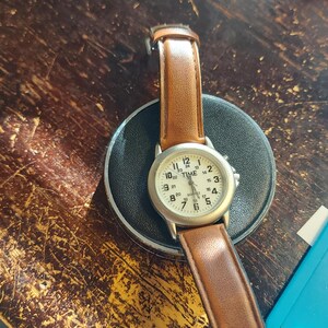 May include: A wristwatch with a light-colored face and brown leather band. The watch face displays the word "TIME" and numerals. The watch is resting on a black circular base, set against a dark, textured wooden surface.