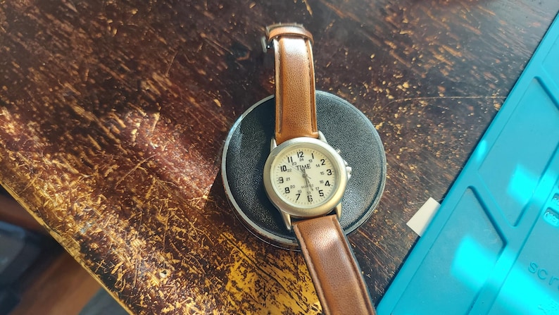 May include: A wristwatch with a light-colored face and brown leather band. The watch face displays the word "TIME" and numerals. The watch is resting on a black circular base, set against a dark, textured wooden surface.
