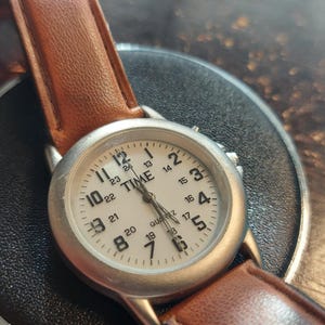 May include: A silver-toned quartz watch with a white face and brown leather strap. The watch face displays the word "TIME" and the numbers 1 through 24. The watch is on a black surface.