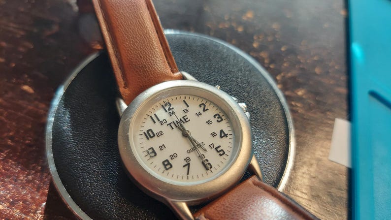 May include: A silver-toned quartz watch with a white face and brown leather strap. The watch face displays the word "TIME" and the numbers 1 through 24. The watch is on a black surface.
