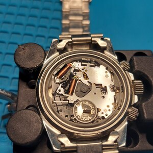 May include: A silver wristwatch, its back removed to show the internal workings. The watch is held in a black watch repair holder on a blue surface. The watch strap is silver. The watch is being repaired.