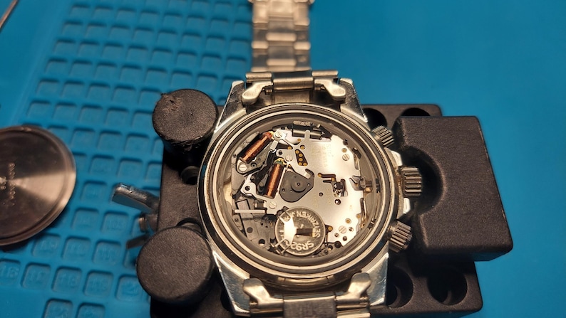 May include: A silver wristwatch, its back removed to show the internal workings. The watch is held in a black watch repair holder on a blue surface. The watch strap is silver. The watch is being repaired.