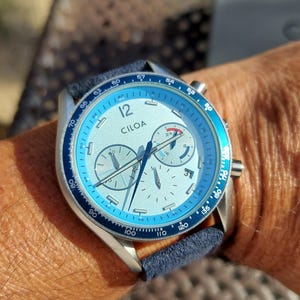 May include: A wristwatch with a light blue bezel and face, featuring the brand name "CILOA". The watch has a silver case and a dark blue band. The watch face includes multiple sub-dials and hands.