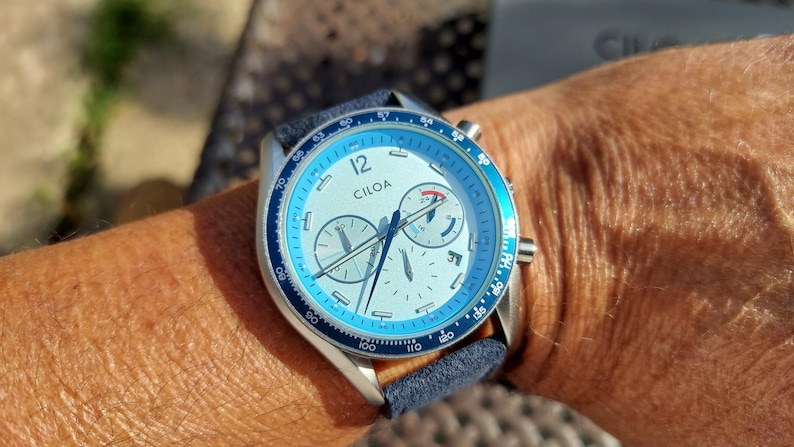 May include: A wristwatch with a light blue bezel and face, featuring the brand name "CILOA". The watch has a silver case and a dark blue band. The watch face includes multiple sub-dials and hands.