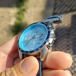 May include: A silver wristwatch with a light blue face and a dark blue bezel. The watch has multiple dials and a gray leather strap. The brand name "CIGA" is visible on the watch face. The watch is held in a hand.