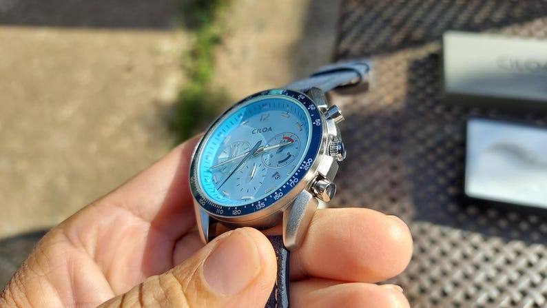 May include: A silver wristwatch with a light blue face and a dark blue bezel. The watch has multiple dials and a gray leather strap. The brand name "CIGA" is visible on the watch face. The watch is held in a hand.
