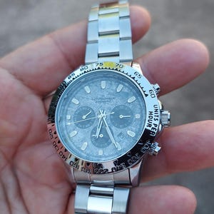 May include: Silver-toned chronograph watch with a textured gray face. The watch features three sub-dials, a rotating bezel with black markings, and a stainless steel band. The watch displays "UNITS PER HOUR" on the bezel.