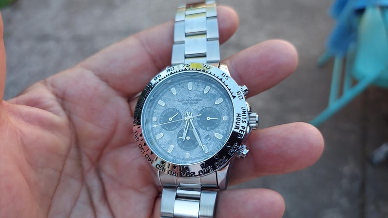 May include: Silver-toned chronograph watch with a textured gray face. The watch features three sub-dials, a rotating bezel with black markings, and a stainless steel band. The watch displays "UNITS PER HOUR" on the bezel.