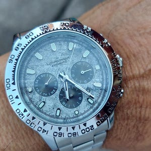May include: A silver wristwatch with a textured gray face and three sub-dials. The watch has a silver bezel with black markings and a silver band. The watch face has the text "LUDWIGSBOURG AUTOMATIC".