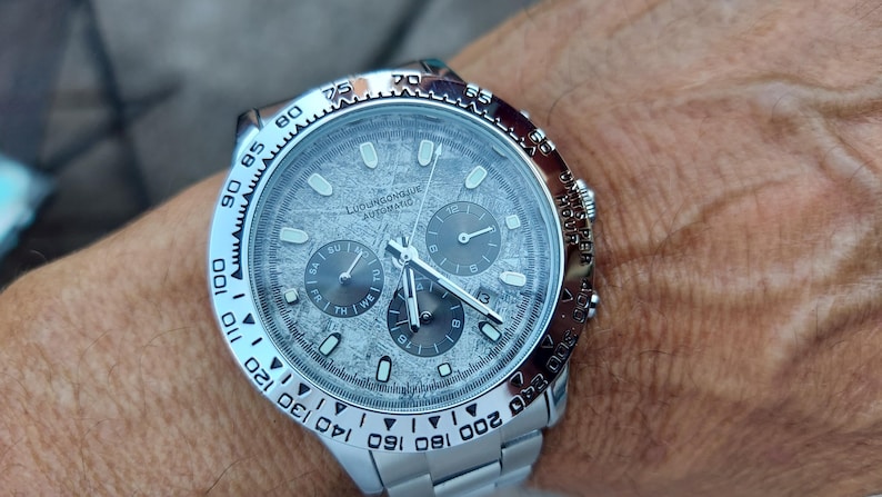May include: A silver wristwatch with a textured gray face and three sub-dials. The watch has a silver bezel with black markings and a silver band. The watch face has the text "LUDWIGSBOURG AUTOMATIC".