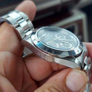 May include: A silver Invicta watch with a black dial. The watch has a stainless steel strap and a round face with multiple sub-dials. The watch is held in a hand.