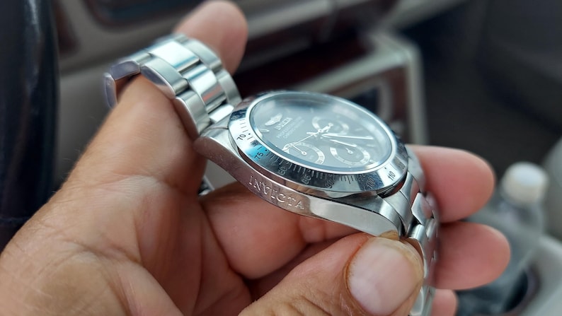 May include: A silver Invicta watch with a black dial. The watch has a stainless steel strap and a round face with multiple sub-dials. The watch is held in a hand.