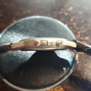 May include: A side view of a wristwatch with a gold-colored case and a dark brown leather strap. The watch face is round, and the strap is attached to the case with small pins. The watch is resting on a dark surface.