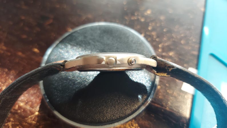 May include: A side view of a wristwatch with a gold-colored case and a dark brown leather strap. The watch face is round, and the strap is attached to the case with small pins. The watch is resting on a dark surface.