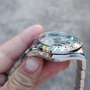 May include: A silver wristwatch with a white dial and a black bezel featuring numbers and markings. The watch has a silver band and is held in a hand.