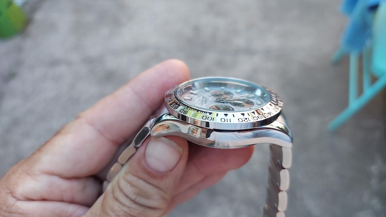May include: A silver wristwatch with a white dial and a black bezel featuring numbers and markings. The watch has a silver band and is held in a hand.