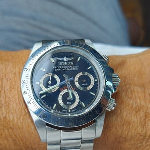 May include: A silver Invicta Professional 200M Chrono-Master watch with a dark blue face. The watch has three sub-dials, a date display, and a silver bezel with white markings. The watch is on a silver metal band.