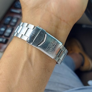 May include: A silver Invicta watch strap is clasped on a wrist. The watch strap is made of linked metal segments. The Invicta logo is visible on the clasp. The person is in a car, with the steering wheel and interior visible.