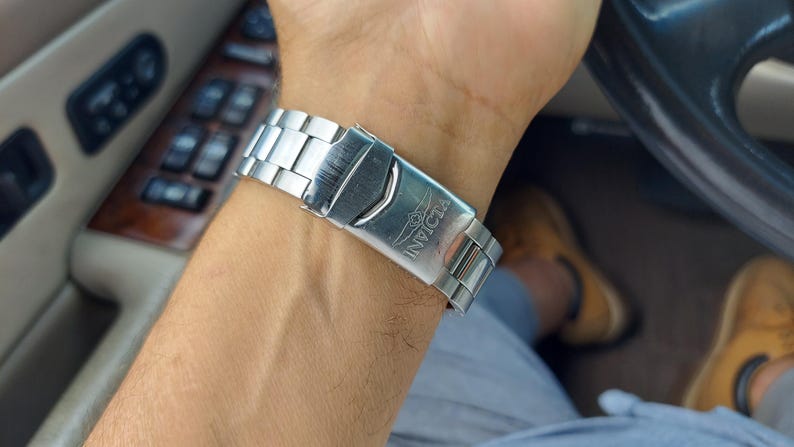 May include: A silver Invicta watch strap is clasped on a wrist. The watch strap is made of linked metal segments. The Invicta logo is visible on the clasp. The person is in a car, with the steering wheel and interior visible.