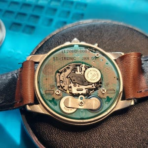 May include: A disassembled wristwatch with a black leather strap and a brown leather accent. The watch face is open, revealing the internal mechanisms, including a battery and circuit board. The watch case is silver-toned.