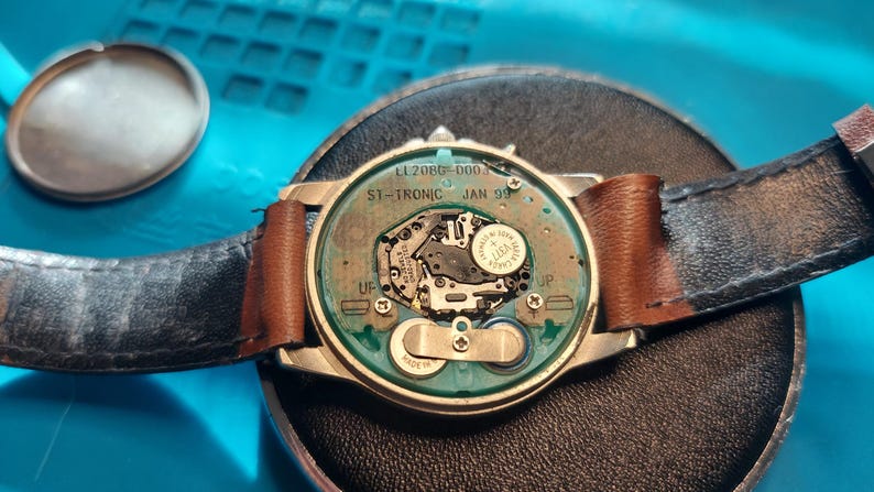 May include: A disassembled wristwatch with a black leather strap and a brown leather accent. The watch face is open, revealing the internal mechanisms, including a battery and circuit board. The watch case is silver-toned.
