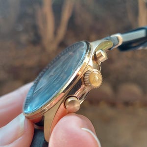May include: A gold-toned wristwatch with a black leather band. The watch face is dark with a light blue hue. The watch has gold-colored buttons and a crown. The watch is held by a person.