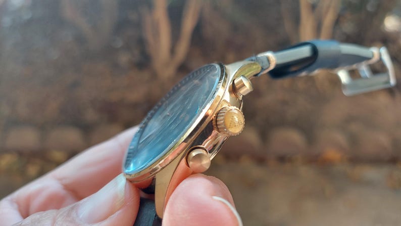 May include: A gold-toned wristwatch with a black leather band. The watch face is dark with a light blue hue. The watch has gold-colored buttons and a crown. The watch is held by a person.