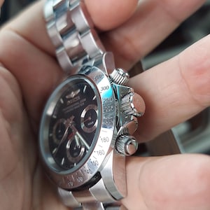 May include: A silver wristwatch with a black face and several sub-dials. The watch has a stainless steel strap and a detailed bezel with numerical markings. The watch is held in a hand, highlighting its design and craftsmanship.
