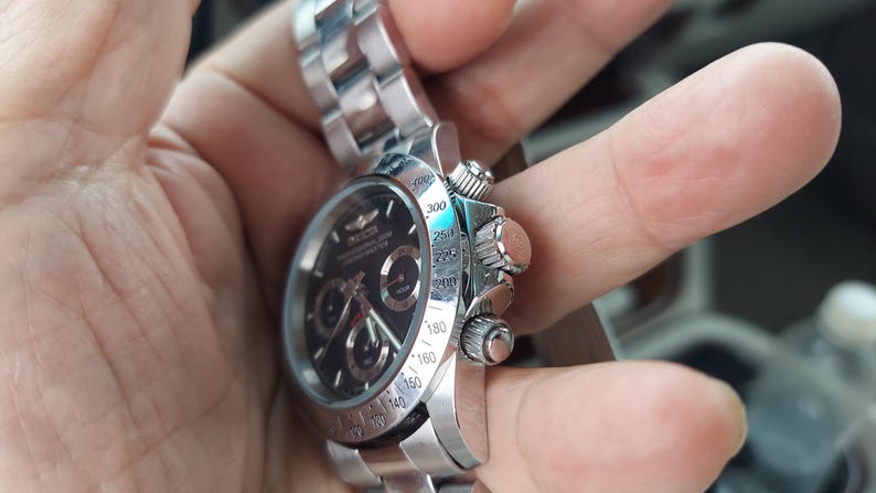 May include: A silver wristwatch with a black face and several sub-dials. The watch has a stainless steel strap and a detailed bezel with numerical markings. The watch is held in a hand, highlighting its design and craftsmanship.