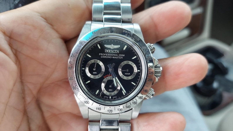 May include: A stainless steel Invicta Professional 200M Chrono-Master watch with a black dial. The watch features three sub-dials, a rotating bezel, and a stainless steel band. The watch has a diameter of approximately 4.5 cm.