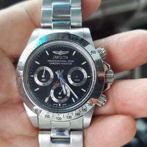 May include: A stainless steel Invicta Chrono-Master watch with a black dial. The watch has three sub-dials, a date display, and a rotating bezel. The watch is held in a hand.