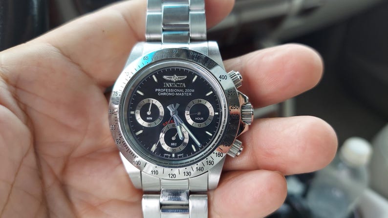 May include: A stainless steel Invicta Chrono-Master watch with a black dial. The watch has three sub-dials, a date display, and a rotating bezel. The watch is held in a hand.