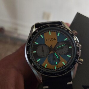 Pu&ograve; includere: Un orologio cronografo CILOA con quadrante nero e oro dotato di scala tachimetrica sulla lunetta. Il quadrante include tre sotto-quadranti, indici luminosi e lancette che si illuminano al buio. Presenta un cinturino nero e una cassa argentata.