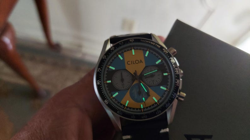 Pu&ograve; includere: Un orologio cronografo CILOA con quadrante nero e oro dotato di scala tachimetrica sulla lunetta. Il quadrante include tre sotto-quadranti, indici luminosi e lancette che si illuminano al buio. Presenta un cinturino nero e una cassa argentata.