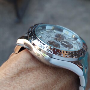 May include: Close-up of a silver wristwatch with a textured gray face and three sub-dials. The watch has a silver bezel with black markings and a white band. The watch is on a person's wrist.