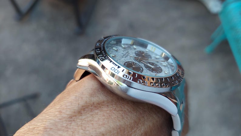 May include: Close-up of a silver wristwatch with a textured gray face and three sub-dials. The watch has a silver bezel with black markings and a white band. The watch is on a person's wrist.