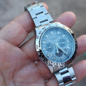 May include: A stainless steel wristwatch with a silver dial and three sub-dials. The watch has a black bezel with white numerals and a silver link bracelet. The watch is held in a hand.