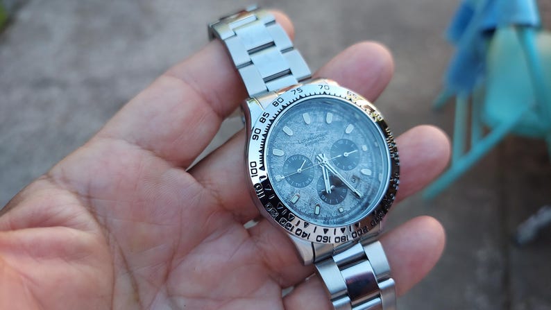 May include: A stainless steel wristwatch with a silver dial and three sub-dials. The watch has a black bezel with white numerals and a silver link bracelet. The watch is held in a hand.