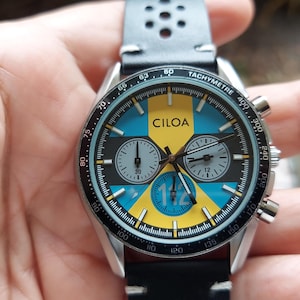Pu&ograve; includere: Un orologio cronografo CILOA con quadrante nero, giallo e blu, tre sotto-quadranti e una scala tachimetrica sulla lunetta. L'orologio presenta un cinturino in pelle nera con dettagli traforati e una cassa in metallo color argento.