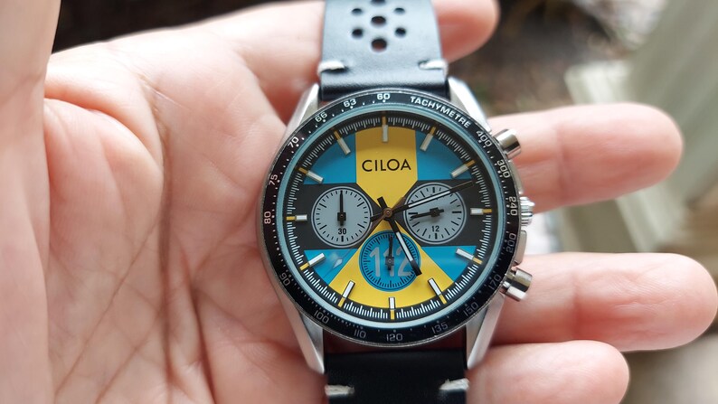 Pu&ograve; includere: Un orologio cronografo CILOA con quadrante nero, giallo e blu, tre sotto-quadranti e una scala tachimetrica sulla lunetta. L'orologio presenta un cinturino in pelle nera con dettagli traforati e una cassa in metallo color argento.
