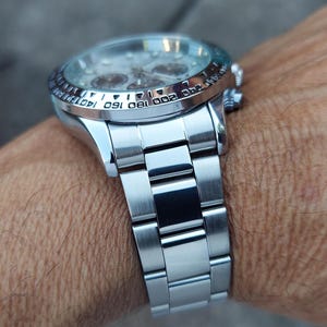 May include: A silver wristwatch with a stainless steel band is shown on a wrist. The watch has a round face with multiple dials and a rotating bezel with numbers. The band is made of rectangular links.